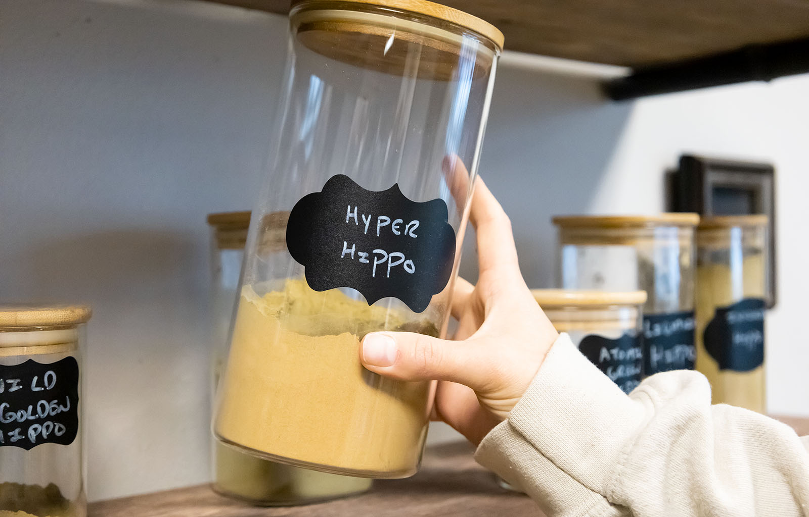 Featured image depicting a shelf containing several jars of maeng da kratom powder. A hand is removing a jar of Green Maeng Da (Hyper Hippo) kratom powder.