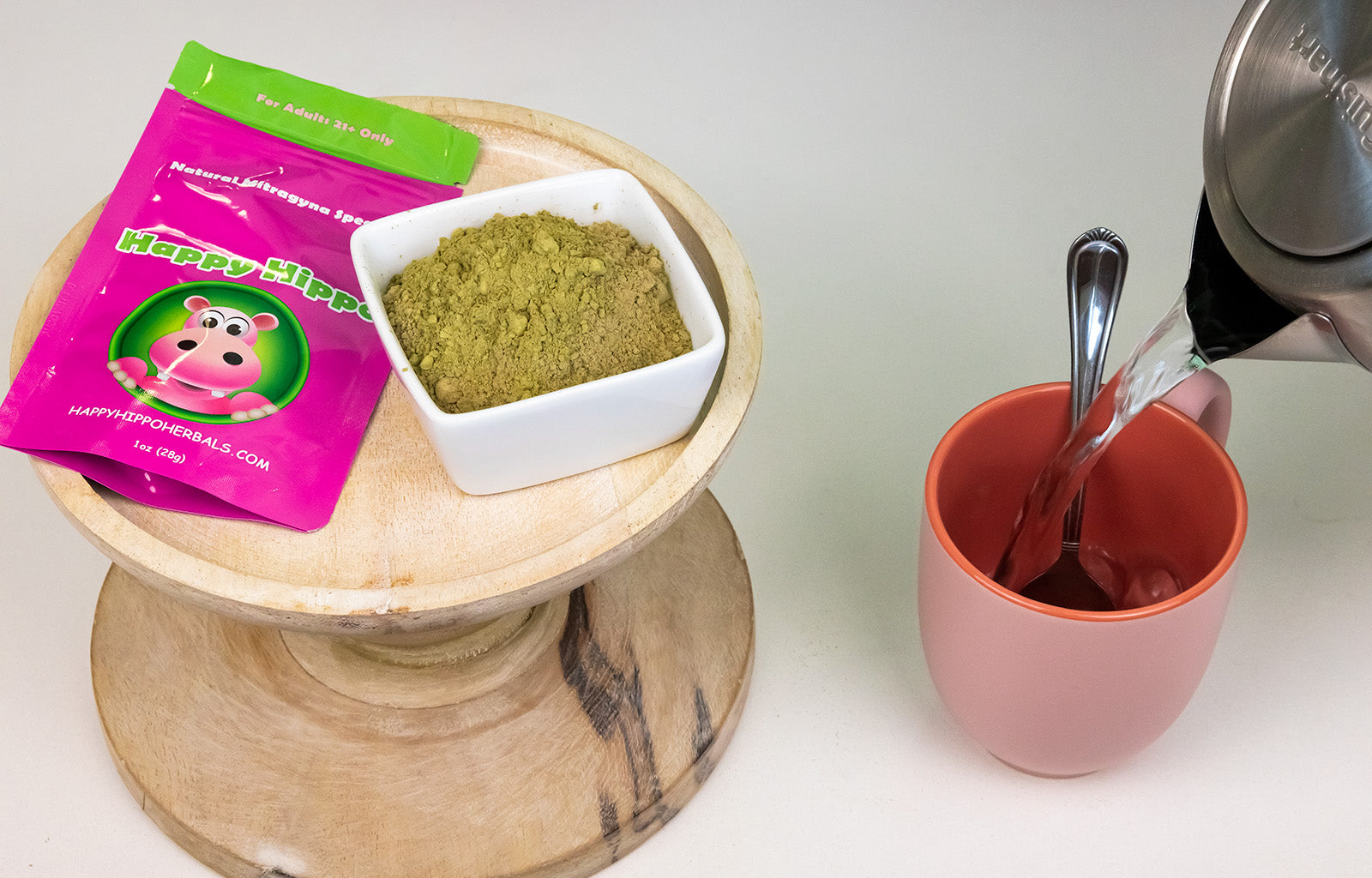 Featured image depicting a small wooden table, atop which sits a packet of Happy Hippo brand White Borneo Kratom Powder. Next to the small wooden table, is a pink teacup being filled with hot water from a steel kettle.