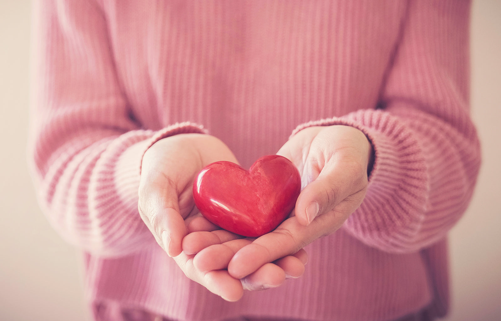 Featured Image depicting a woman in a pink sweater, holding a large red heart in her outstretched hands. The image is representative of Kratom Heart Health.