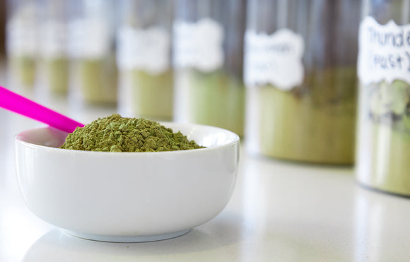 Featured image depicting a row of jars filled with various kratom strains, in front of which is a clean white bowl heaped with Kratom Powder and a 1-gram little pink measuring scoop.