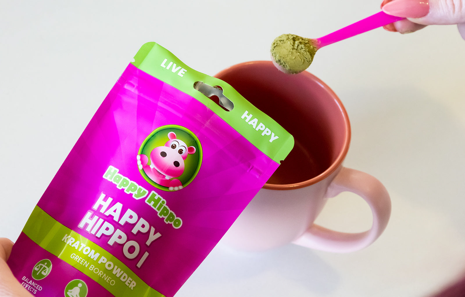 Photographic image depicting a pink coffee cup, over which someone is using a little pink measuring scoop adding Happy Hippo brand kratom powder to their morning tea.