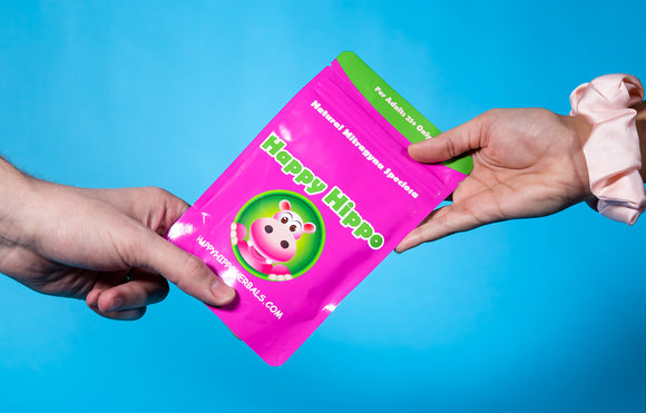 Featured image depicting the hand-off of a 1oz packet of Happy Hippo brand Green Maeng Da kratom powder. The hand-off is from a man's hand, to a woman's hand against a blue background.